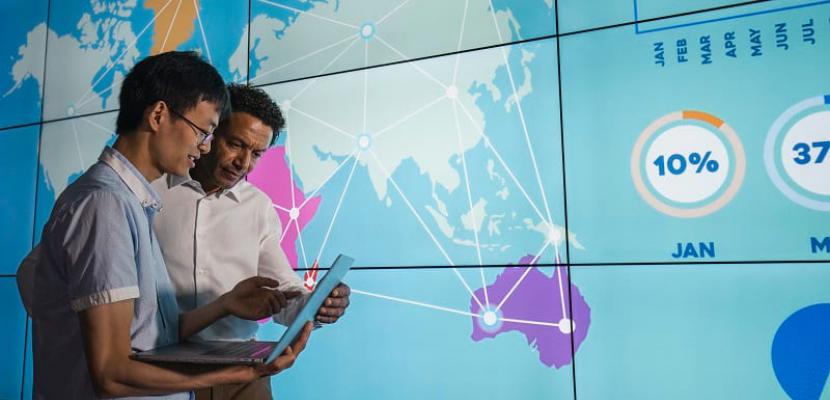 Two men look at a projection of a map chart on a wall